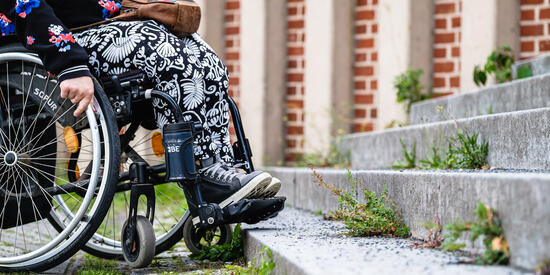 Beine einer Frau, die im Rollstuhl sitzt. Sie steht vor einer steilen Treppe.