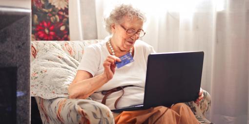 Ältere Dame im Sessel sitzend beim Online-Shopping, Laptop auf dem Schoß und Kreditkarte in der Hand.