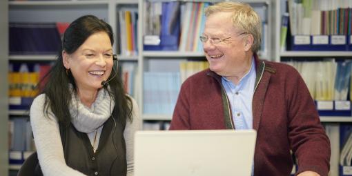 Frau mit Kopfhörer und Mann sitzen lachend vor einem Laptop