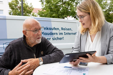 Zwei Personen sitzen an einem Tisch. Die jüngere Person hat ein Tabelt in der Hand und dreht iden Bildschirm zur ältern Person.