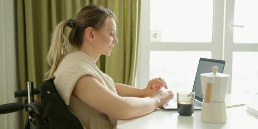 Eine junge Frau sitzt am Tisch in einem Rollstuhl. Sie nutzt einen Laptop. 