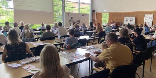 Im Konferenzsaal sitzen Personen an den Tischen. Sie sind von hinten zu sehen. Vorn eine große Leinwand mit Referent.