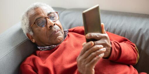 Ein älterer Herr liegt auf dem Sofa. In der Hand hat er ein Smartphone, auf das er schaut.
