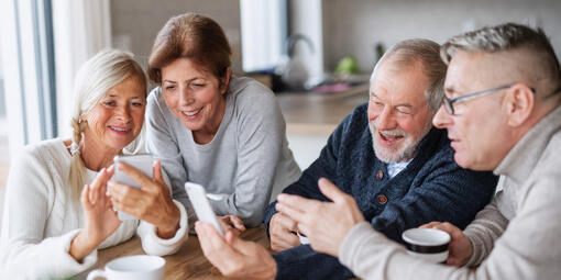 Zwei Frauen und zwei Männer sitzen gemeinsam am Tisch. Eine Frau und ein Mann halten jeweils ein Smartphone in der Hand. Die Gruppe schaut fröhlich gemeinsam auf die Geräte.