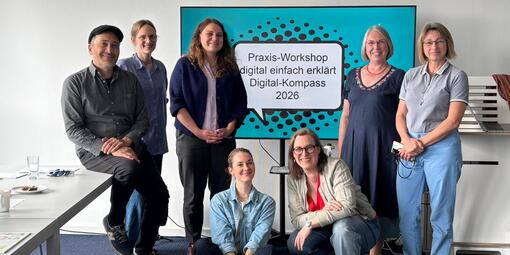 Die Teilnehmerinnen und Teilnehmer der Schulung stehen vor einem Bildschirm. Dort zeigt es die Titelfolie der Präsentation an: "Praxis-Workshop digital einfach erklärt Digital-Kompass 2026". 