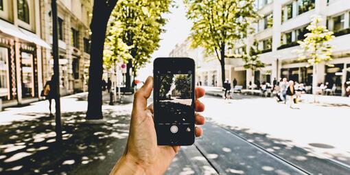 Eine Person ist in einer Stadt unterwegs und hält ein Smartphone. Die Person macht gerade ein Foto der Straße. Es sind Straßenbahnschienen, Gebäude und Bäume zu sehen. 
