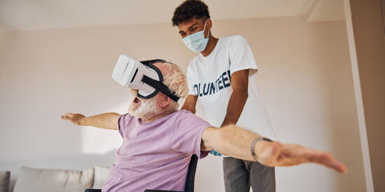 Ein Mann sitzt im Rollstuhl, breitet die Arme zur Seite aus als würde er fliegen und trägt eine VR-Brille. Dahinter ist ein junger Mann mit Maske, der ihn im Rollstuhl schiebt.