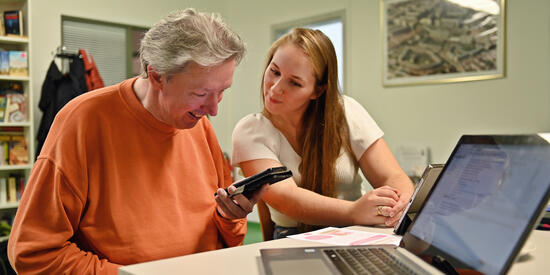 Ein älterer Mann und eine junge Frau schauen gemeinsam auf das Smartphone in der Hand des Mannes. Davor steht ein aufgeklappter Laptop.