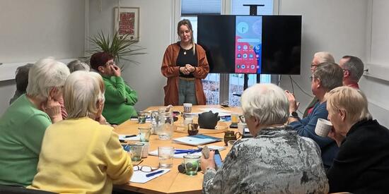 Die Workshop-Leiterin erklärt etwas zur digitalen Welt. Hinter ihr ist ein Bildschirm. Vor ihr sitzen neun Seniorinnen und Senioren.