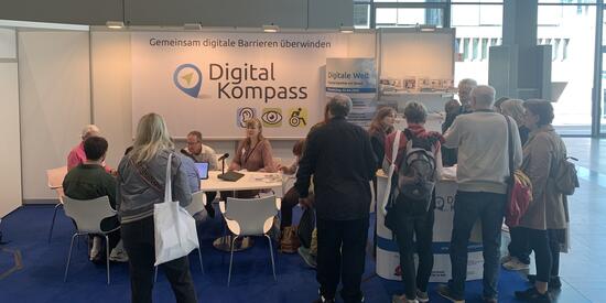 Messestand des Digital-Kompass. Links ein Tisch an dem einige Personen sitzen. Rechts der Messethresen vor dem viele Personen stehen. Im Hintergrund in Großformat das Logo des Digital-Kompass. 