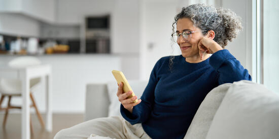 Eine ältere Dame sitzt fröhlich auf dem Sofa und schaut auf ihr Handy.