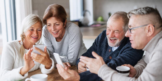 Zwei Frauen und zwei Männer sitzen gemeinsam am Tisch. Eine Frau und ein Mann halten jeweils ein Smartphone in der Hand. Die Gruppe schaut fröhlich gemeinsam auf die Geräte.