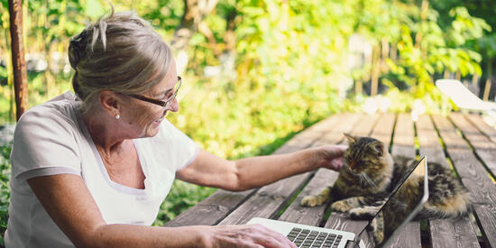 Éine ältere Dame sitzt draußen an einem Holztisch. Sieht nutzt einen Laptop und streichelt dabei eine Katze, die auf dem Tisch liegt.