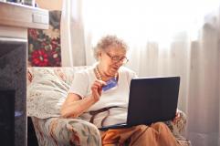Ältere Dame im Sessel sitzend beim Online-Shopping, Laptop auf dem Schoß und Kreditkarte in der Hand.