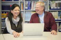 Frau mit Kopfhörer und Mann sitzen lachend vor einem Laptop