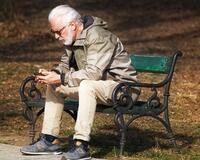 Ein älterer Herr sitzt mit seinem Smartphone in der Hand auf einer Parkband.