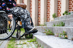 Beine einer Frau, die im Rollstuhl sitzt. Sie steht vor einer steilen Treppe.