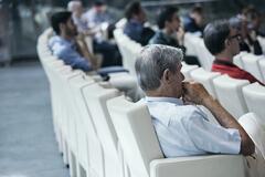 Konferenzraum mit weißen Stühlen und einigen Personen. Im Vordergrund ist ein älterer Mann, der aufmerksam zuhört.