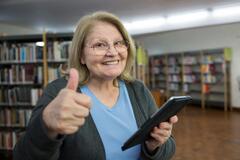 Eine ältere Dame hält ein Tablet in der Hand und zeigt einen Daumen nach oben. Sie steht vermutlich in einer Bibliothek und hat ein breites Grinsen. 