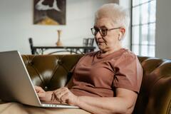 Eine ältere Dame sitzt auf dem Sofa. Sie trägt Kopfhörer und nutzt ihren Laptop.