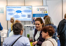Messestand des Digital-Kompass. Im Hintergrund ein Bildschirm sowie ein Aufsteller des Projektes. Davor viele Personen im Austausch.
