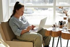 Eine junge Frau mit Down-Syndrom sitzt auf dem Sofa. Sie trägt Kopfhörer und nutzt einen Laptop auf ihrem Schoß.