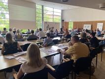 Im Konferenzsaal sitzen Personen an den Tischen. Sie sind von hinten zu sehen. Vorn eine große Leinwand mit Referent. 
