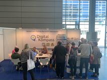 Messestand des Digital-Kompass. Links ein Tisch an dem einige Personen sitzen. Rechts der Messethresen vor dem viele Personen stehen. Im Hintergrund in Großformat das Logo des Digital-Kompass. 