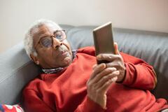 Ein älterer Herr liegt auf dem Sofa. In der Hand hat er ein Smartphone, auf das er schaut.