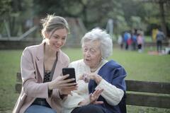 Eine junge Frau und eine ältere Dame sitzen auf einer Parkbank. Die jüngere Frau hält ein Smartphone in der Hand und zeigt der älteren Dame grinsend etwas. Die ältere Dame sieht interessiert aus und zeigt auf das Smartphone.