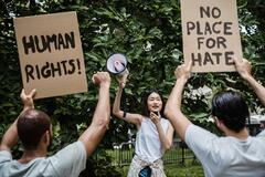 Eine Frau hält ein Megafon in die Luft und spricht in ein Aufnahmegerät. Vor ihr stehen zwei Männer, ihr Rücken ist zu sehen. Sie halten Schilder hoch. Eines mit der Schrift "Humans Rights", das andere mit "No place for hate".
