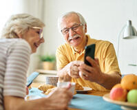 Zwei ältere Personen sitzen am Tisch. Der ältere Herr zeigt der älteren Dame etwas auf dem Handy und beide schauen fröhlich.