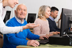 Ein Computerkabinett, in dem mehrere Personen sitzen. Im Vordergrund ist ein älterer Herr der zu einer Dame schaut, die ihm etwas am PC erklärt.