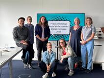Die Teilnehmerinnen und Teilnehmer der Schulung stehen vor einem Bildschirm. Dort zeigt es die Titelfolie der Präsentation an: "Praxis-Workshop digital einfach erklärt Digital-Kompass 2026". 