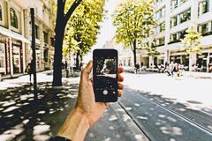 Eine Person ist in einer Stadt unterwegs und hält ein Smartphone. Die Person macht gerade ein Foto der Straße. Es sind Straßenbahnschienen, Gebäude und Bäume zu sehen. 