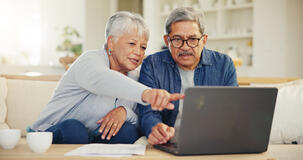 Zwei ältere Personen sitzen auf der Couch vor einem Laptop. Die Frau zeigt mit dem Finger auf den Laptop.