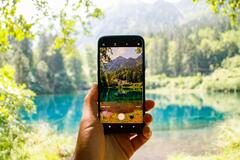 Ein Handy fotografiert einen blauen See im Wald. Im Vordergrund das Smartphone mit der Aufnahme, im Hintergrund die besagte Szene.