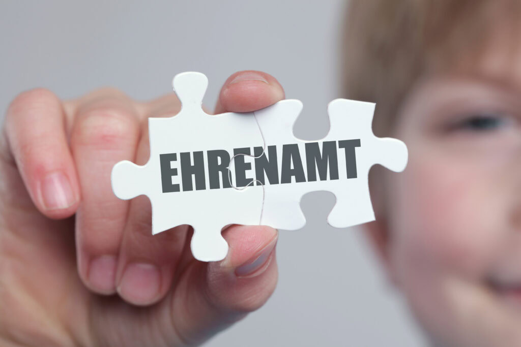 Ein Puzzlestück auf dem "Ehrenamt" steht wird gezeigt. Im Hintergrund mit halben Gesicht ist eine Person zu sehen.