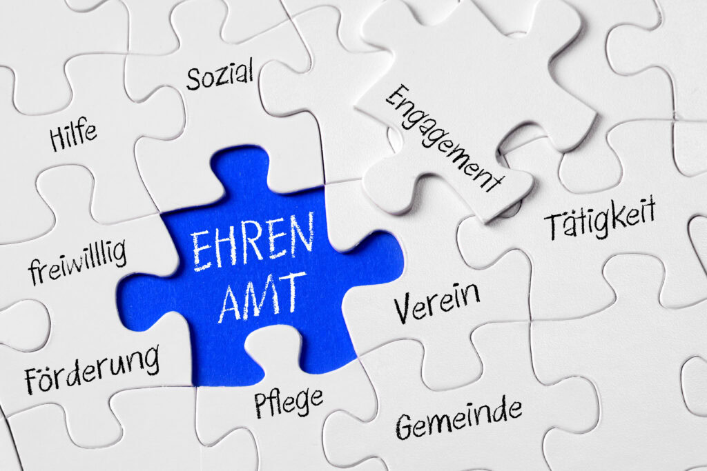 Ein Puzzle, in dem ein Teil fehlt. Diese Lücke ist blau dargestellt und ihr steht "Ehrenamt" auf den anderen Puzzleteilen drum herum steht beispielsweise Hilfe, Pflege oder Gemeinde. Diese Puzzleteile sind weiß.