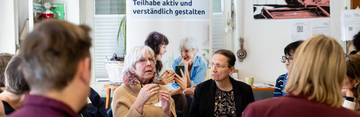 Mehrere Personen sitzen in einem hellen Raum in einer Gesprächsrunde. Im Mittelpunkt spricht eine Person und gestikuliert mit den Händen, während andere Teilnehmende zuhören. Im Hintergrund ist ein Roll-up-Banner mit dem Text „Teilhabe aktiv und verständlich gestalten“ sowie ein Foto von Personen im Gespräch zu sehen.
