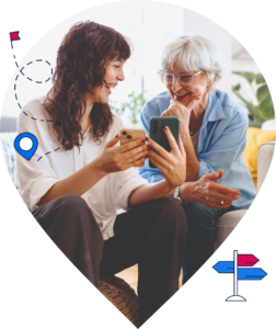 Eine jüngere und eine ältere Frau sitzen gemeinsam auf einer Couch. Die jüngere Frau zeigt der älteren Frau etwas auf zwei Handy-Bildschirmen, beide lächeln.