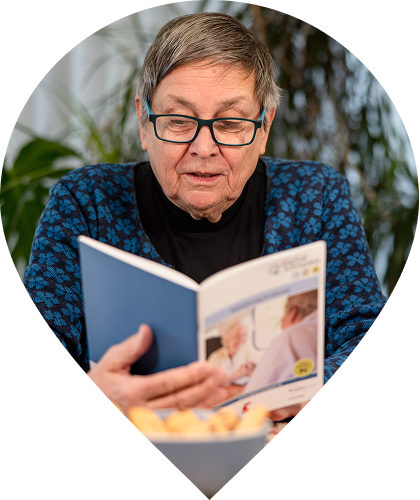Eine Person mit Brille sitzt an einem Tisch und liest eine Broschüre mit blauem Umschlag. Auf dem Cover sind Informationsgrafiken und ein Foto abgebildet. Im Vordergrund steht eine Schale mit Gebäck, im Hintergrund sind Zimmerpflanzen in einem hellen Innenraum zu sehen.