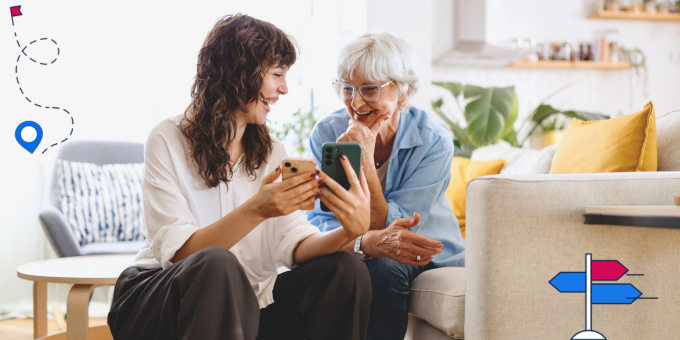 Eine jüngere und eine ältere Frau sitzen gemeinsam auf einer Couch. Die jüngere Frau zeigt der älteren Frau etwas auf zwei Handy-Bildschirmen, beide lächeln.