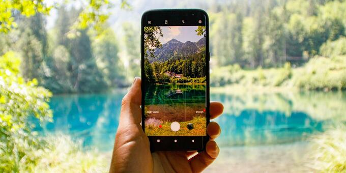 im Hintergrund ist ein See mitten im Grünen. Im Vordergrund ist eine Hand, die ein Smartphone hält und ein Foto vom See macht.